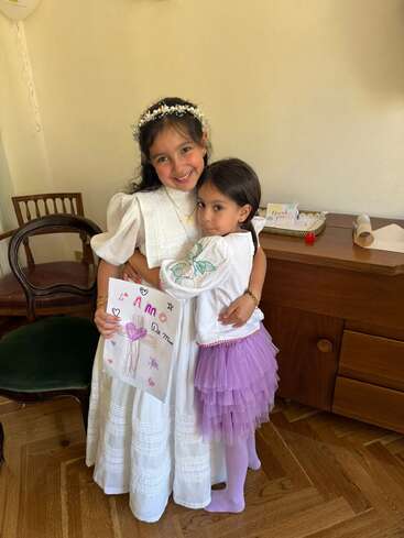 Zwei junge Mädchen umarmen sich herzlich in einem Raum. Das ältere Mädchen trägt ein weißes Kleid und einen Blumenkranz und hält eine bunte Zeichnung in der Hand. Das jüngere trägt ein lila Tutu und lächelt sanft.