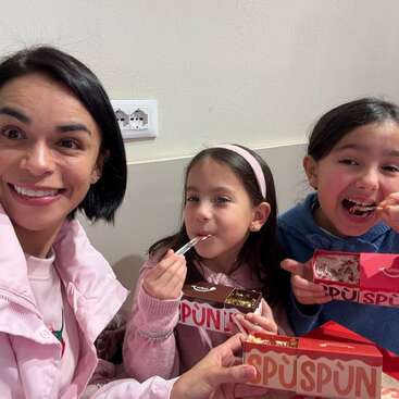A woman and two young girls happily enjoy eating desserts together. They are smiling, wearing cozy clothes, and holding red and white dessert boxes labeled “SPÜN.”