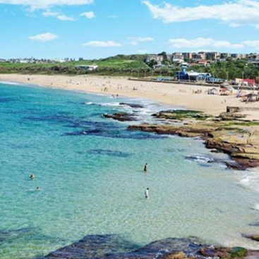 The image depicts a serene beach scene with a sandy shore, clear blue water, and a rocky shoreline, set against a backdrop of buildings and lush greenery.