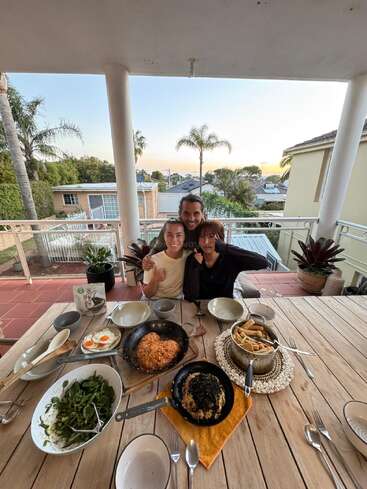 Trois personnes sourient à une table dressée en plein air pour le dîner. Différents plats délicieux sont servis, entourés de verdure luxuriante et de palmiers, capturant un moment joyeux.