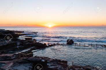 L'image représente une scène sereine de l'océan au lever ou au coucher du soleil, avec un rivage rocheux, de douces vagues et un ciel vibrant aux teintes orange et jaune.