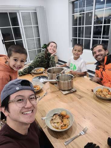 L'image montre une famille de cinq personnes assises autour d'une table avec de la nourriture, souriant à l'appareil photo, dans une pièce lumineuse aux murs blancs et aux grandes fenêtres.