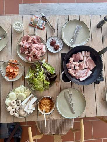 Une table en bois dressée pour un barbecue coréen avec du porc cru, des légumes verts, des champignons, des oignons, du kimchi, des sauces, des nouilles instantanées et une poêle grésillant avec de la viande.