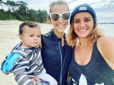 L'image montre deux femmes et un enfant sur une plage, la femme de gauche tenant l'enfant et l'autre femme portant un bonnet et un débardeur.