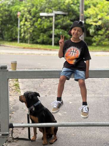 Un garçon souriant, vêtu d'un maillot de basket et d'une casquette, est assis sur un rail et affiche un signe de paix. En dessous, un chien noir et brun attend patiemment.
