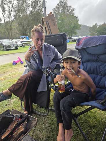 Una mujer hace el signo de la paz sentada junto a un niño sin camiseta que sostiene un biberón y un palo, en un camping con coches, barcos y árboles al fondo.