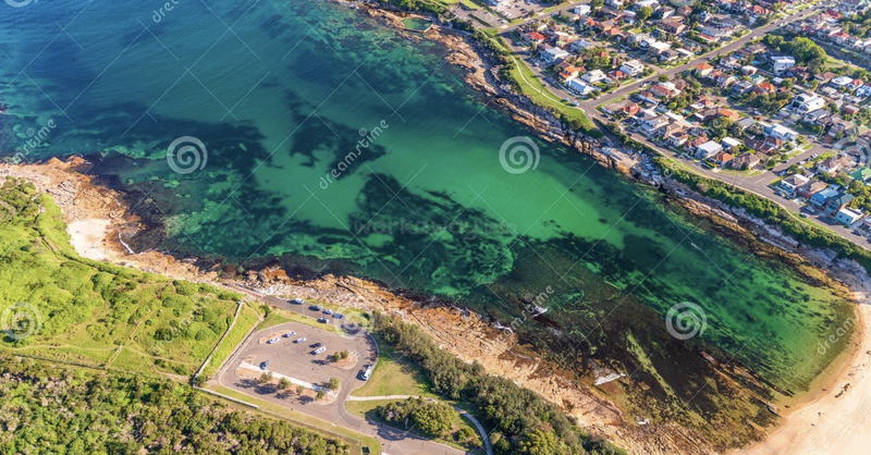 Vue aérienne d'une côte turquoise vibrante avec des rivages rocheux, un parking, des pelouses vertes, des voitures éparpillées et des maisons résidentielles à proximité, mêlant magnifiquement la terre et la mer.