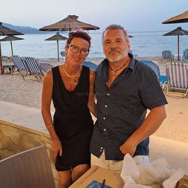 A smiling couple poses at a beachside restaurant, with straw umbrellas, loungers, and calm sea in the background. Evening light creates a relaxed, vacation atmosphere.