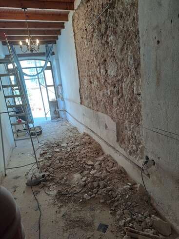 This image shows a room under renovation, with a partially demolished wall, exposed ceiling beams, loose debris on the floor, a ladder, and construction tools.