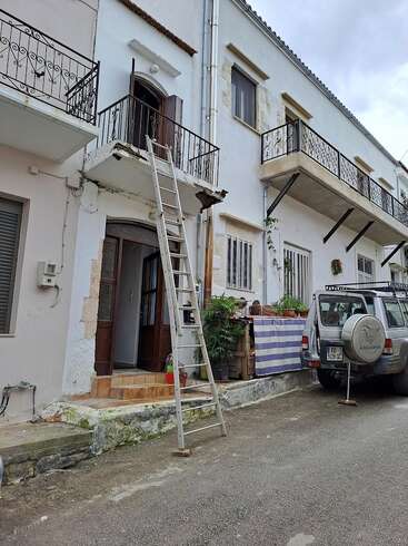 A white building with balconies, a metal ladder leading to an upper door, potted plants, a striped cloth, and a parked SUV on a quiet street.
