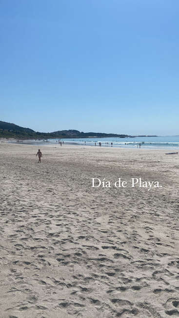 Das Bild zeigt eine ruhige Strandszene mit einer weiten Sandfläche, ein paar Menschen, die am Ufer spazieren gehen, und einen klaren blauen Himmel über sich.
