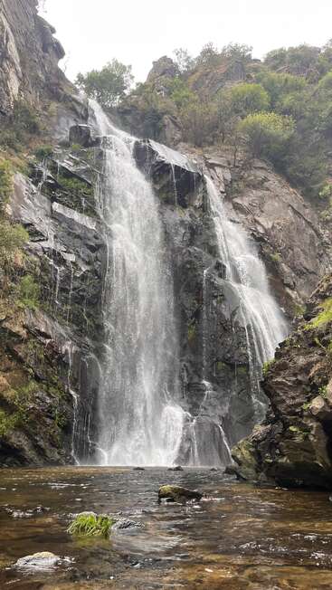 Das Bild zeigt einen majestätischen Wasserfall, der in Kaskaden einen felsigen Abhang hinunterstürzt, umgeben von üppigem Grün, mit einem ruhigen Wasserbecken an seinem Fuß.