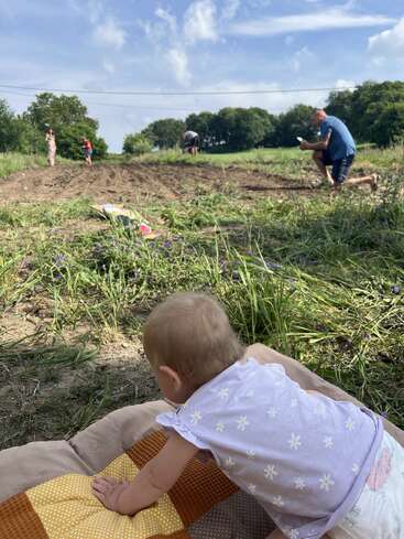 Das Bild zeigt eine ruhige Szene mit einem Baby, das auf einer Decke auf einem Feld liegt, umgeben von Menschen, die im Hintergrund arbeiten, mit Bäumen und blauem Himmel.