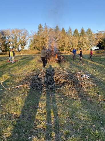 Eine Gruppe von Menschen versammelt sich um ein großes Lagerfeuer auf einer Wiese, umgeben von Bäumen und einem strahlend blauen Himmel, mit einem Haufen Äste in der Nähe.