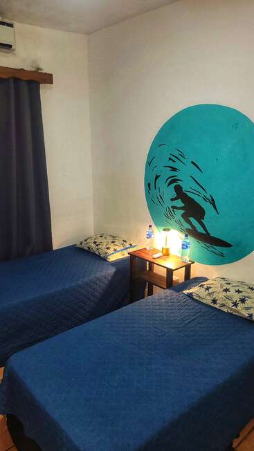 This cozy room features two single beds with blue covers, tropical-themed pillows, a wooden nightstand with lamp, bottled water, and a surfer mural on the wall.