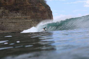 A surfer rides a powerful wave near a rocky cliff, skillfully maneuvering through the water under a blue sky on an adventurous, sunny day by the ocean.