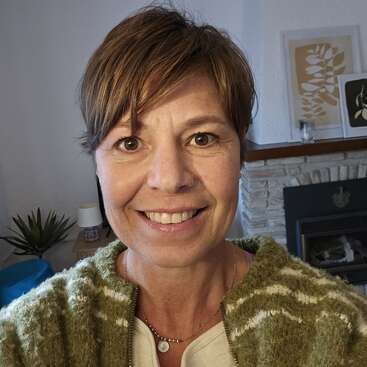 Una mujer sonriente con el pelo castaño corto se hace un selfie en el interior. Lleva un jersey verde. La acogedora habitación tiene obras de arte, una chimenea y una planta.
