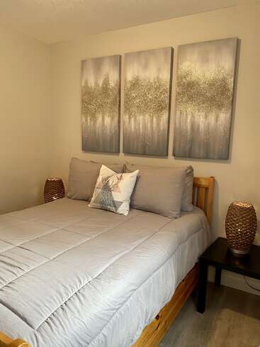 A cozy bedroom with a wooden bed frame, gray bedding, a geometric deer pillow, abstract wall art, two woven lamps, and neutral-colored walls creates a relaxing atmosphere.