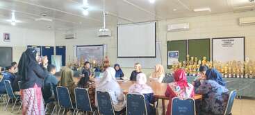 A group of people sit around a conference table in a meeting room, surrounded by numerous trophies. One person stands taking a photo. Bright, formal setting.