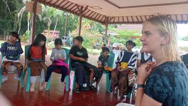 A group of children and one adult sit in a gazebo, engaging in conversation. The setting is tropical, with lush vegetation and traditional attire visible.