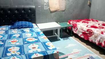 This image shows a small room with two beds, one with a Doraemon bedsheet and pillow, another with a red floral blanket, a simple white table, and gray walls.