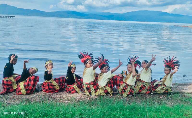 A group of children in traditional costumes sits by the lakeside, smiling and flashing peace signs, with mountains and blue water in the background.