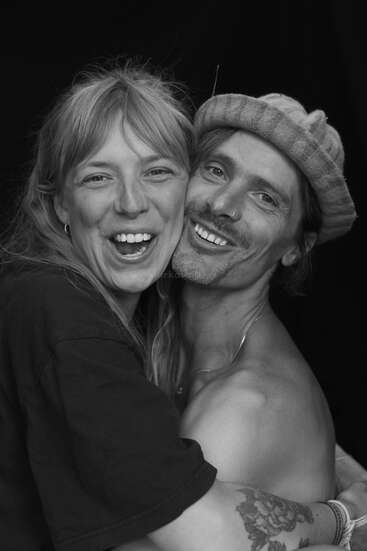 Una pareja alegre se abraza, ambos sonriendo ampliamente. La mujer tiene el pelo claro y lleva una camisa oscura. El hombre lleva gorro, vello facial y un aro en la nariz.