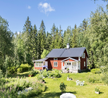 Esta imagen muestra una encantadora casa roja con tejado oscuro, rodeada de exuberante vegetación, altos árboles, un vibrante jardín y asientos al aire libre bajo un cielo despejado y soleado.