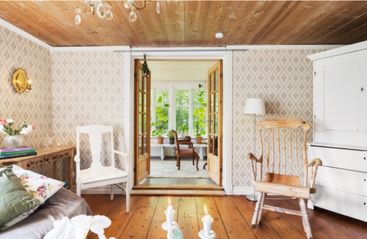 Una acogedora sala de estar rústica con suelos de madera, papel pintado estampado, muebles de madera, detalles florales y luz solar que entra por las puertas de cristal a una luminosa sala de estar.