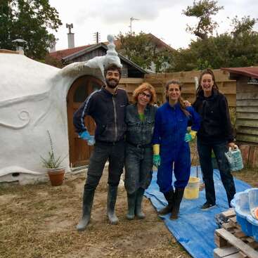 The image shows four people standing in front of a white structure, wearing work clothes and rubber boots, with a blue tarp and buckets nearby, suggesting a construction or renovation project.