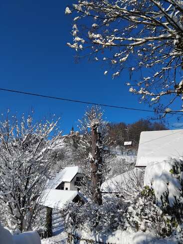 A picturesque winter village scene features snow-covered rooftops, frosty trees, clear blue sky, and a distant church tower nestled among white, sparkling hills and woods.