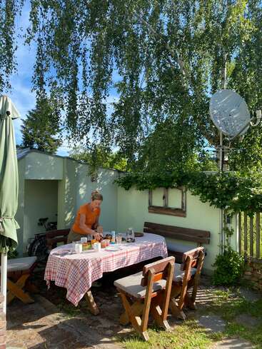 Une femme dresse une table d'extérieur recouverte d'une nappe à carreaux rouges. Entourée de bancs et de verdure, la lumière du soleil filtre à travers les arbres, créant ainsi un jardin d'été confortable.