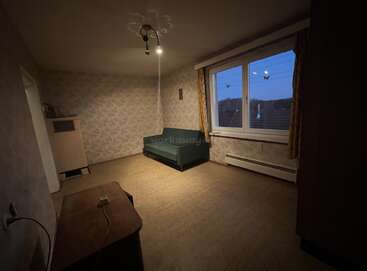 Una habitación poco iluminada y escasamente amueblada con un sofá verde, un escritorio, una gran ventana con cortinas, paredes empapeladas, suelo enmoquetado y una única lámpara de techo.