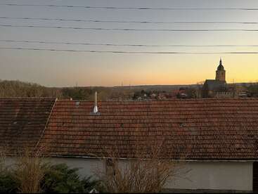 A tranquil village scene at sunset, featuring red-tiled rooftops, silhouetted trees, a church with a steeple, distant hills, and power lines above. Peaceful ambiance.