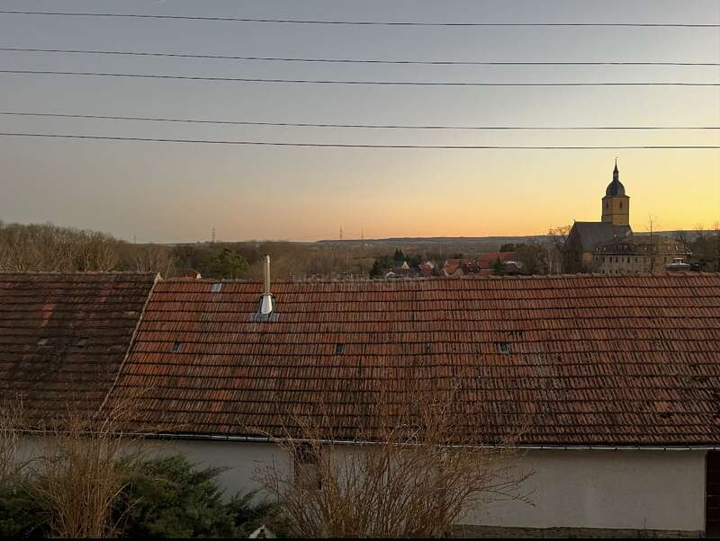 Tranquila escena de un pueblo al atardecer, con tejados de tejas rojas, árboles en silueta, una iglesia con campanario, colinas lejanas y líneas eléctricas por encima. Ambiente tranquilo.