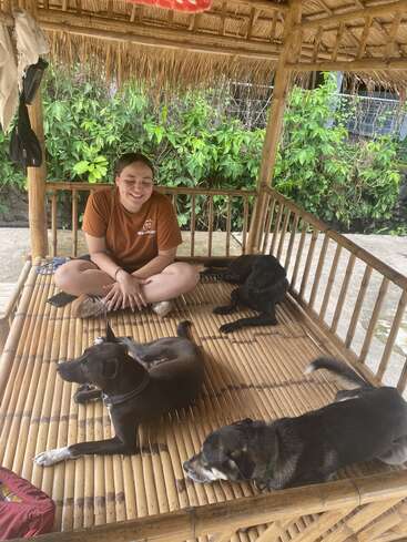 Una persona sonriente está sentada con las piernas cruzadas en la plataforma de una cabaña de bambú, rodeada de tres perros relajados. Las exuberantes plantas verdes y el rústico techo de paja crean un entorno tranquilo y natural.