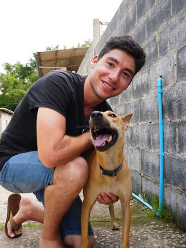 Un joven sonriente está agachado junto a un perro feliz de color marrón claro. Están al aire libre junto a una pared de ladrillo gris, ambos parecen alegres y relajados juntos.