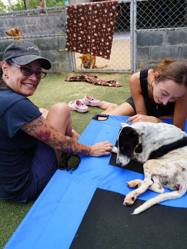 Dos mujeres sonrientes sentadas en una colchoneta azul cuidan con delicadeza a un perro blanco y negro. Al fondo se ve otro perro y los alrededores del refugio.