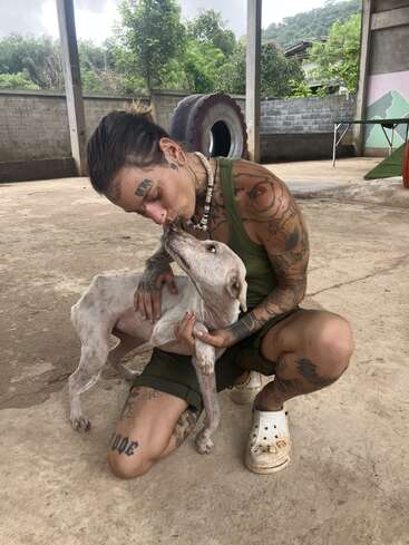 Una persona tatuada se agacha, abraza y besa cariñosamente a un perro blanco y delgado. Están al aire libre, rodeados de árboles, hormigón y un gran neumático al fondo.