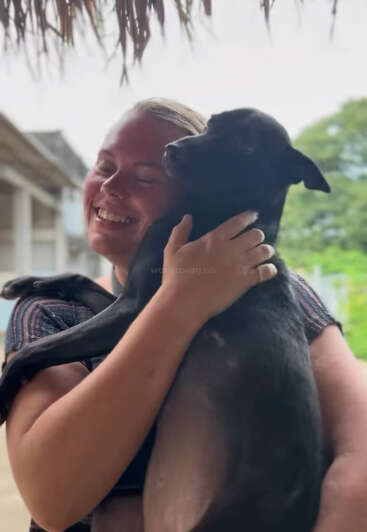 Una mujer abraza alegremente a un perro negro, ambos parecen felices y contentos. Están al aire libre bajo un tejado de paja con vegetación y edificios al fondo.