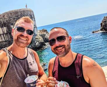 Two men wearing sunglasses and tank tops smile for a selfie by the sea. They’re enjoying ice cream with a historic stone fortress and blue water behind them.