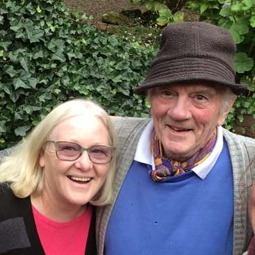 La imagen muestra a un hombre y una mujer mayores posando para una foto, con la mujer a la izquierda y el hombre a la derecha, ambos sonriendo a la cámara.