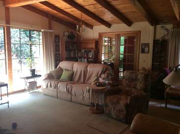 La imagen muestra un acogedor salón con un sofá, un sillón y una estantería, con vigas de madera en el techo y una gran ventana con vistas a los árboles del exterior.