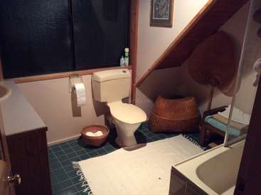 La imagen muestra un cuarto de baño con inodoro, lavabo, bañera y varios accesorios, con una alfombra blanca en el suelo y un armario de madera contra la pared.