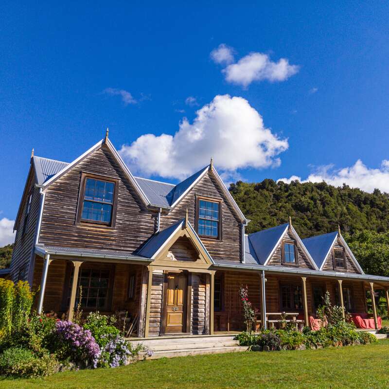 A rustic wooden house with a tin roof stands under a bright blue sky. Surrounded by lush greenery and colorful flowers, it exudes countryside charm.
