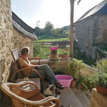 The image depicts a serene scene of a man sitting on a porch with a dog, surrounded by potted plants and a picturesque rural landscape in the background.