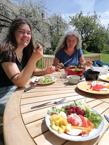 L'image représente deux femmes prenant un repas sur une table de patio en bois, entourées d'un jardin serein avec des arbres en fleurs et un ciel bleu.