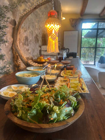 A rustic wooden table holds a fresh salad, eggs, dips, and vegetables under a warm, decorative lamp in a cozy, inviting, stone-walled dining area.