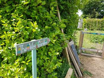Un cartel de madera con la inscripción «The Veg Garden» (El huerto) se encuentra junto a unos frondosos arbustos verdes, señalando un camino cerrado que conduce a un huerto con plantas en crecimiento y herramientas de jardinería cerca.