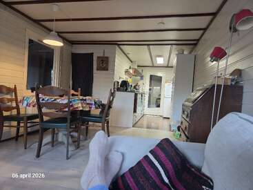 Un salon chaleureux avec des murs en bois, une table à manger, une cuisine en arrière-plan et une personne qui se détend sur un canapé, chaussée de chaussettes et enveloppée dans une couverture.